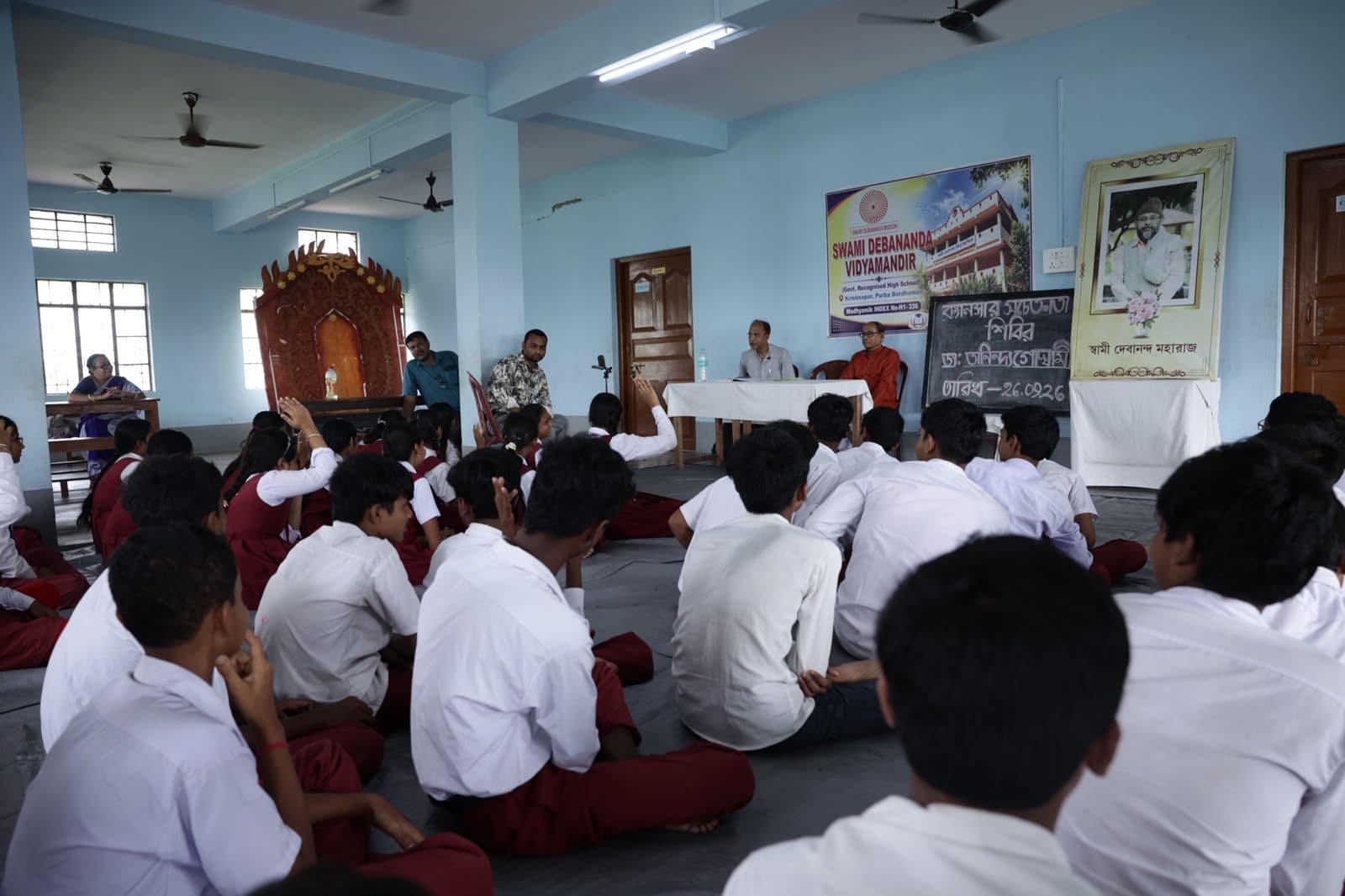 Students seated on the floor of a hall, seen from behind, facing a speaker's table and a chalkboard
