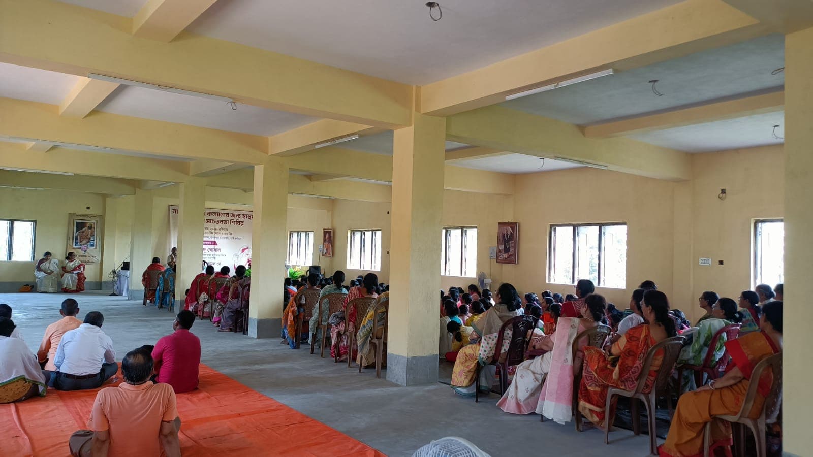 A long pillared hall with women seated on chairs and on mats along its sides