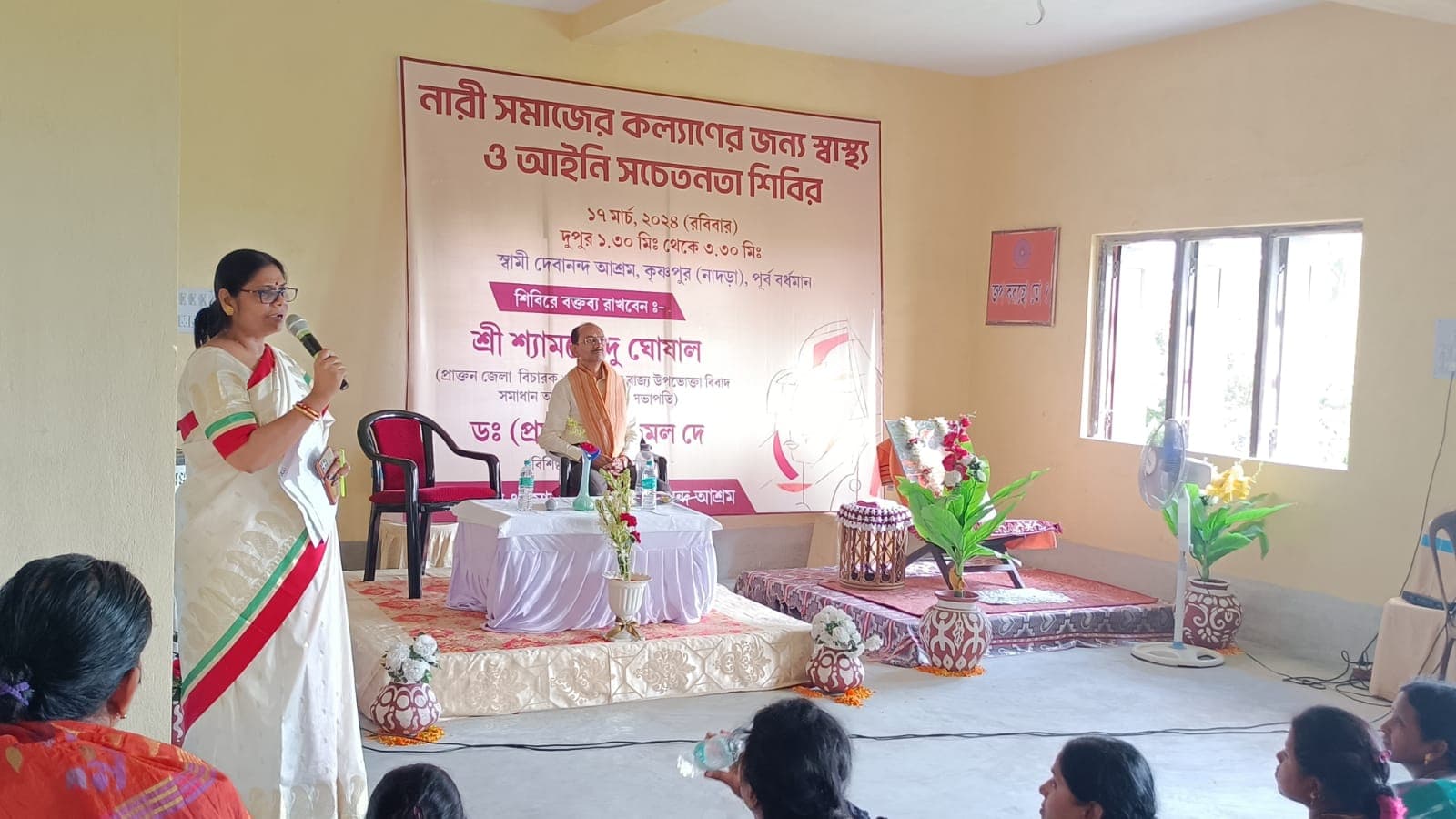 A woman speaking into a microphone to a seated audience, in front of a banner lettered in Bengali