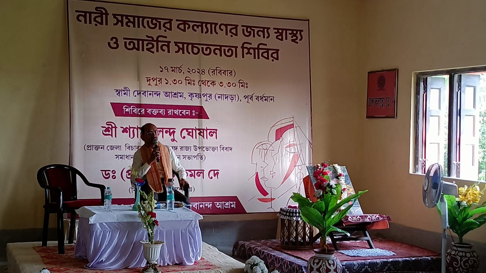 A speaker's table with flowers on a dais, beneath a large banner lettered in Bengali