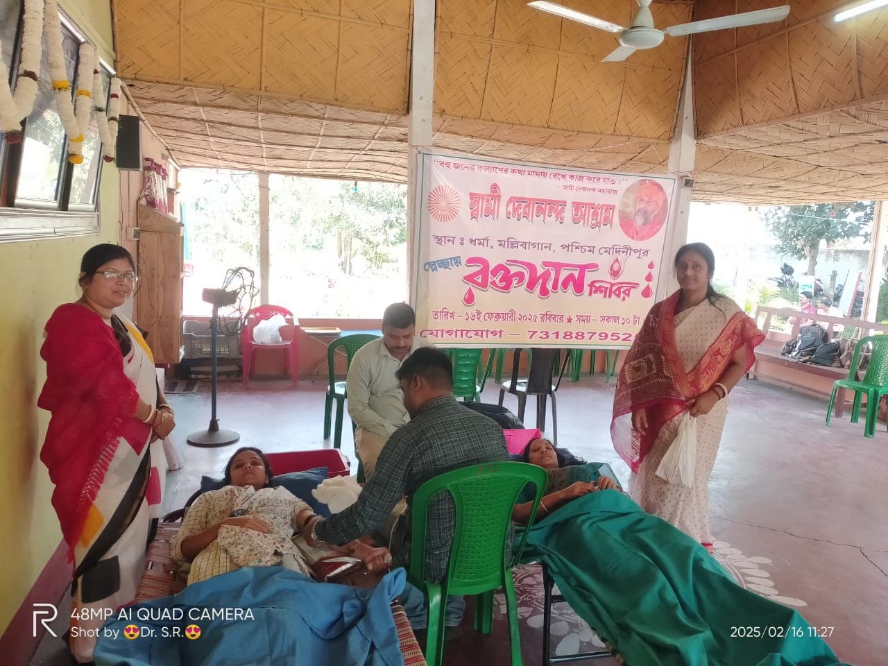 A blood donation camp: donors resting on beds in a hall, with a banner on the wall