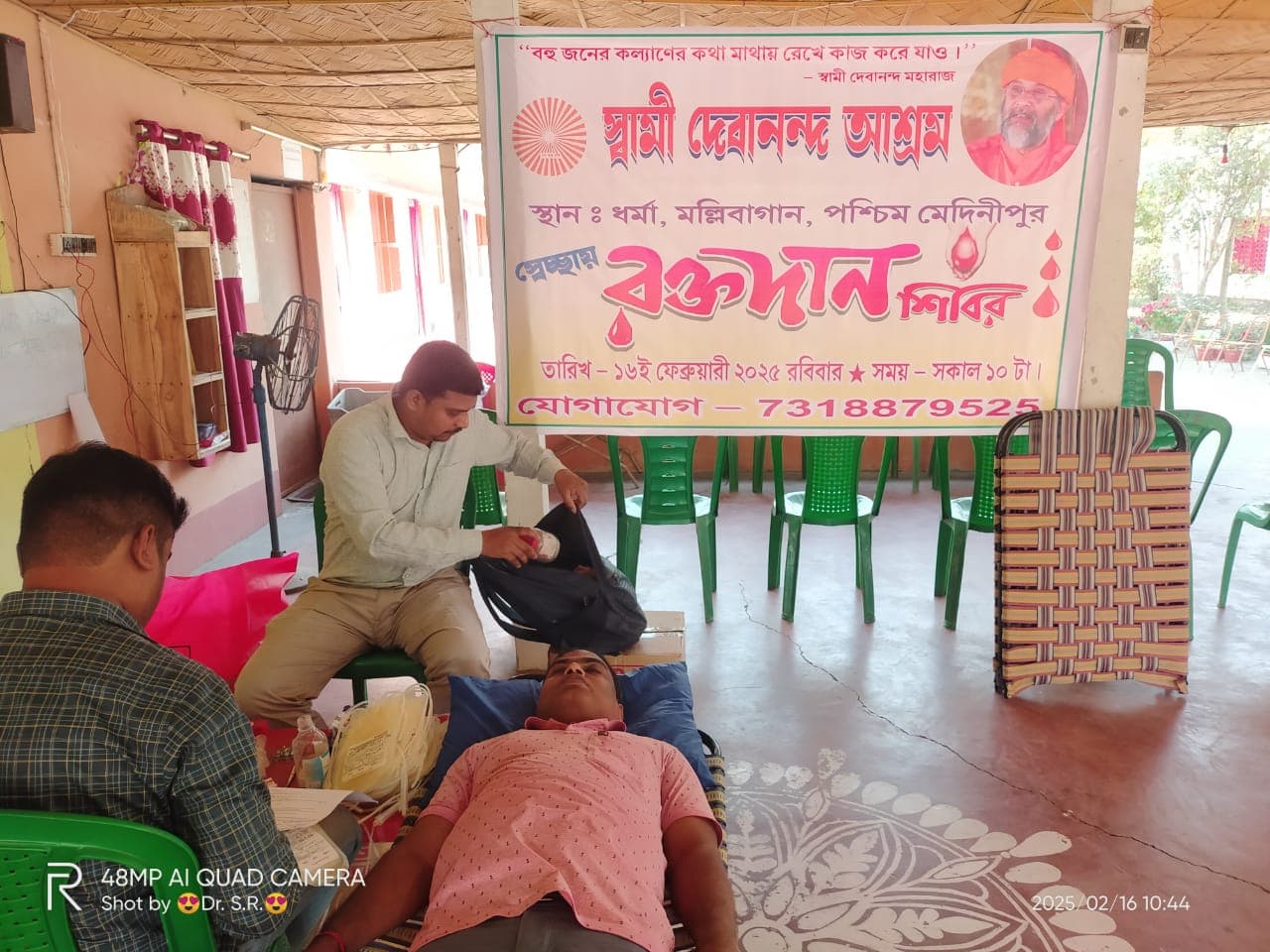 A blood donation camp: donors resting on beds in a hall, with a banner on the wall