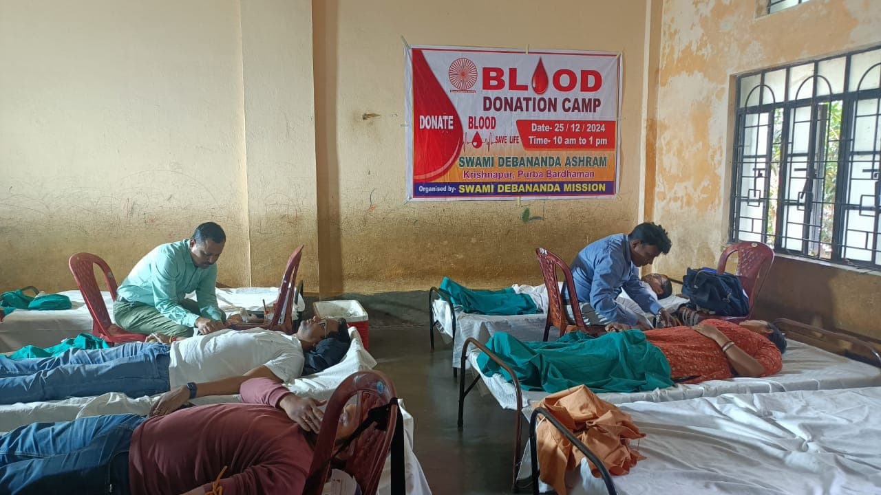 A blood donation camp: donors resting on beds in a hall, with a banner on the wall