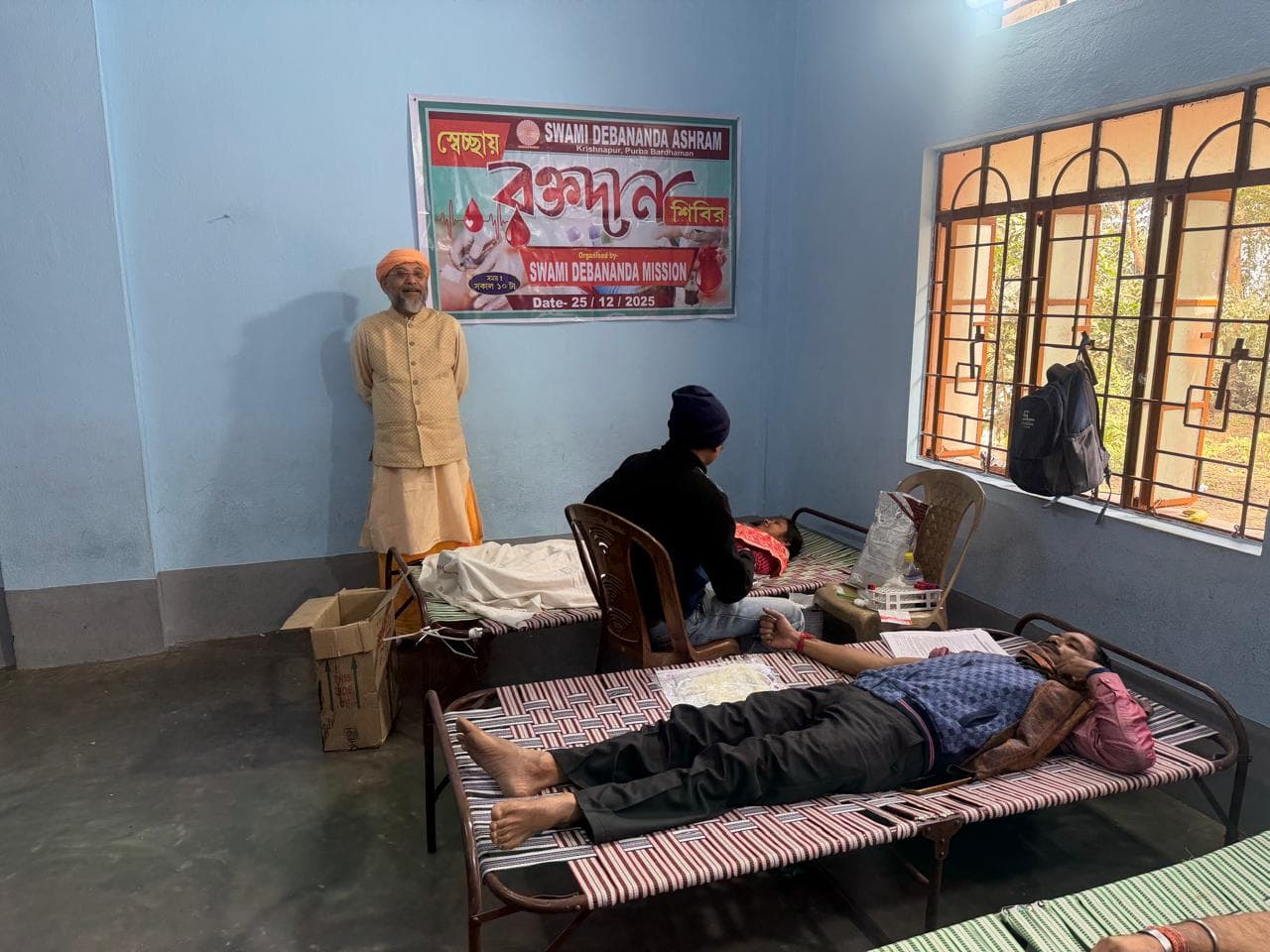 A blood donation camp: donors resting on beds in a hall, with a banner on the wall