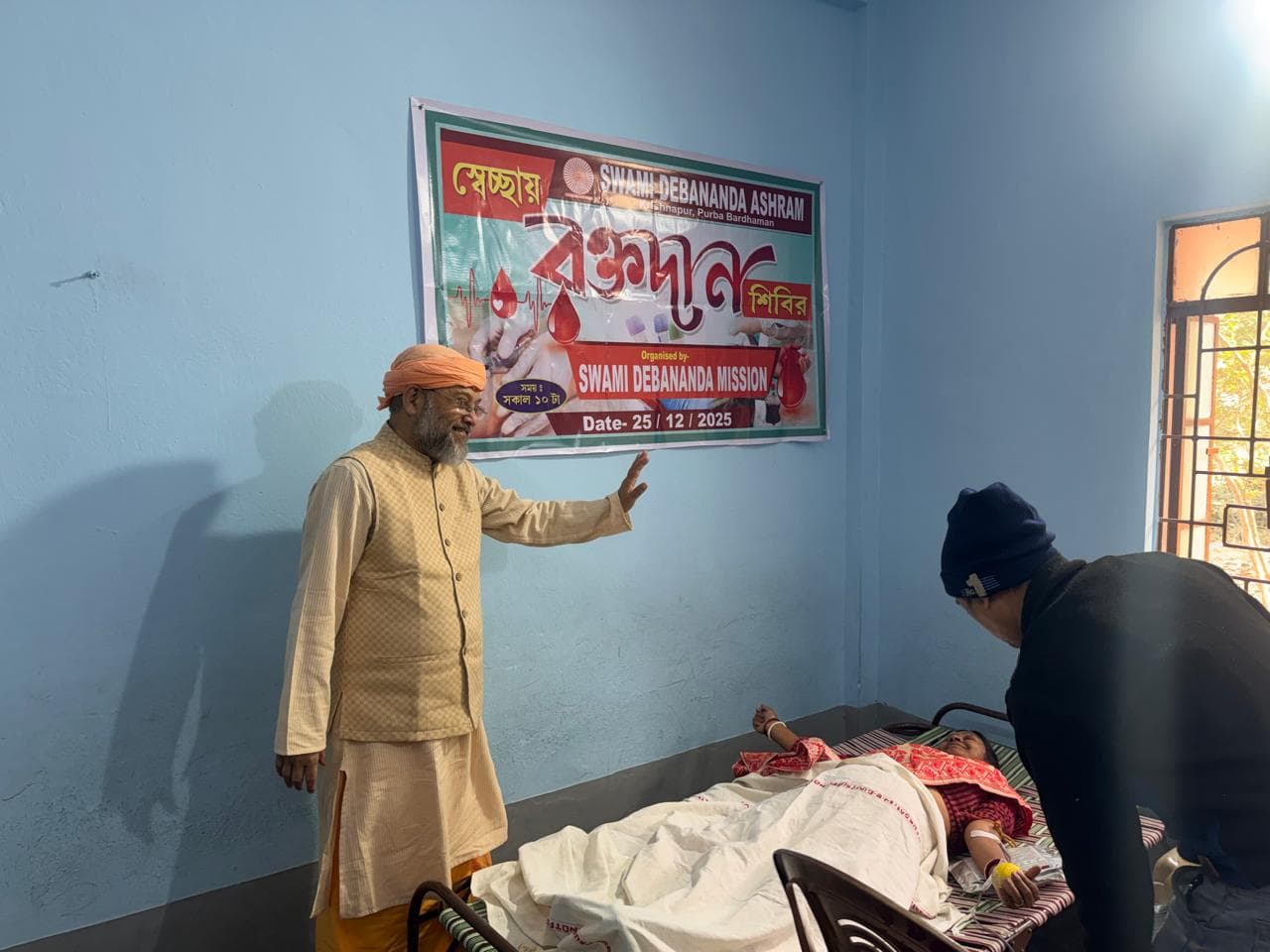 A blood donation camp: donors resting on beds in a hall, with a banner on the wall