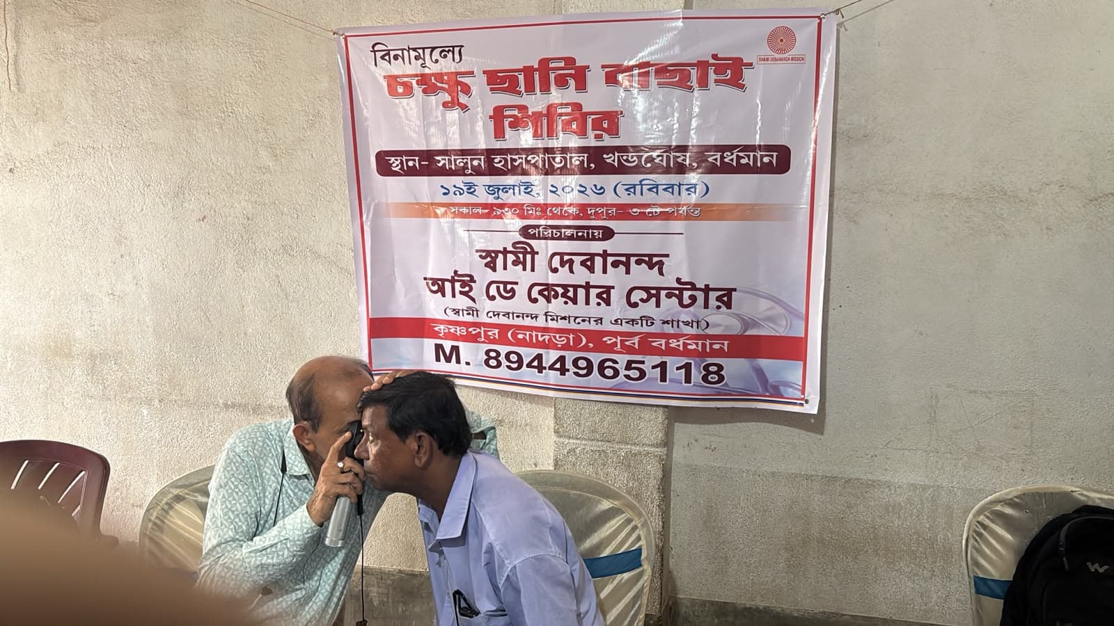 An eye screening camp: a doctor seeing a patient at a table, with a banner lettered in Bengali behind