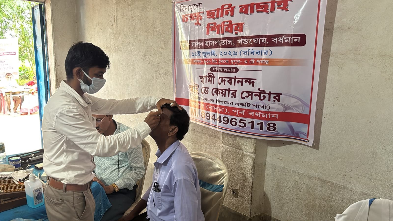 An eye screening camp: a doctor seeing a patient at a table, with a banner lettered in Bengali behind
