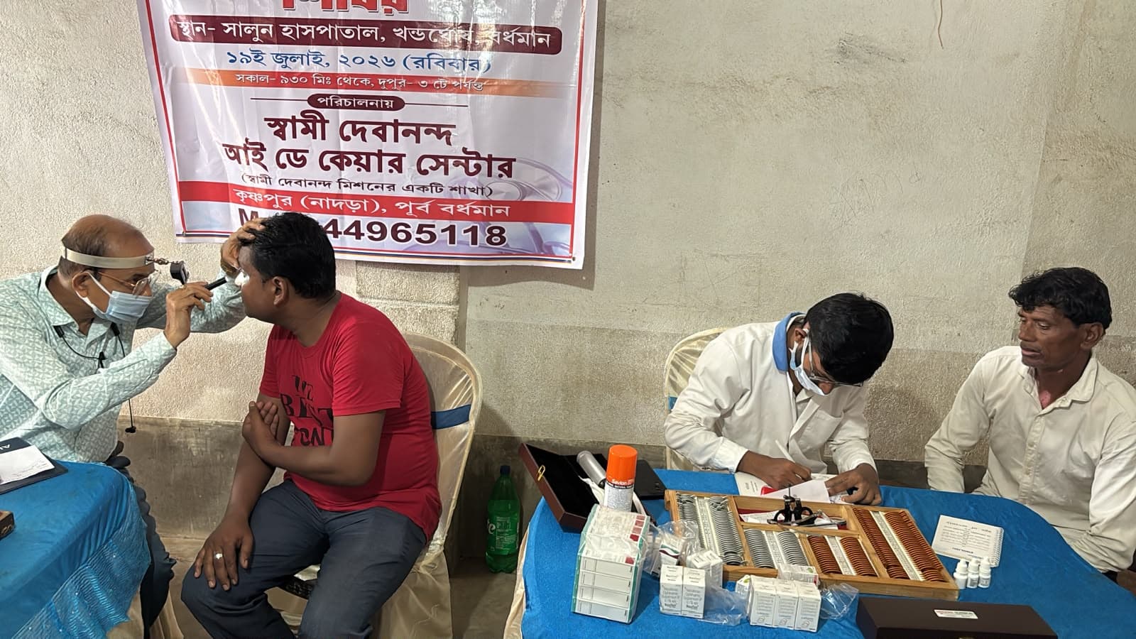 An eye screening camp: a doctor seeing a patient at a table, with a banner lettered in Bengali behind