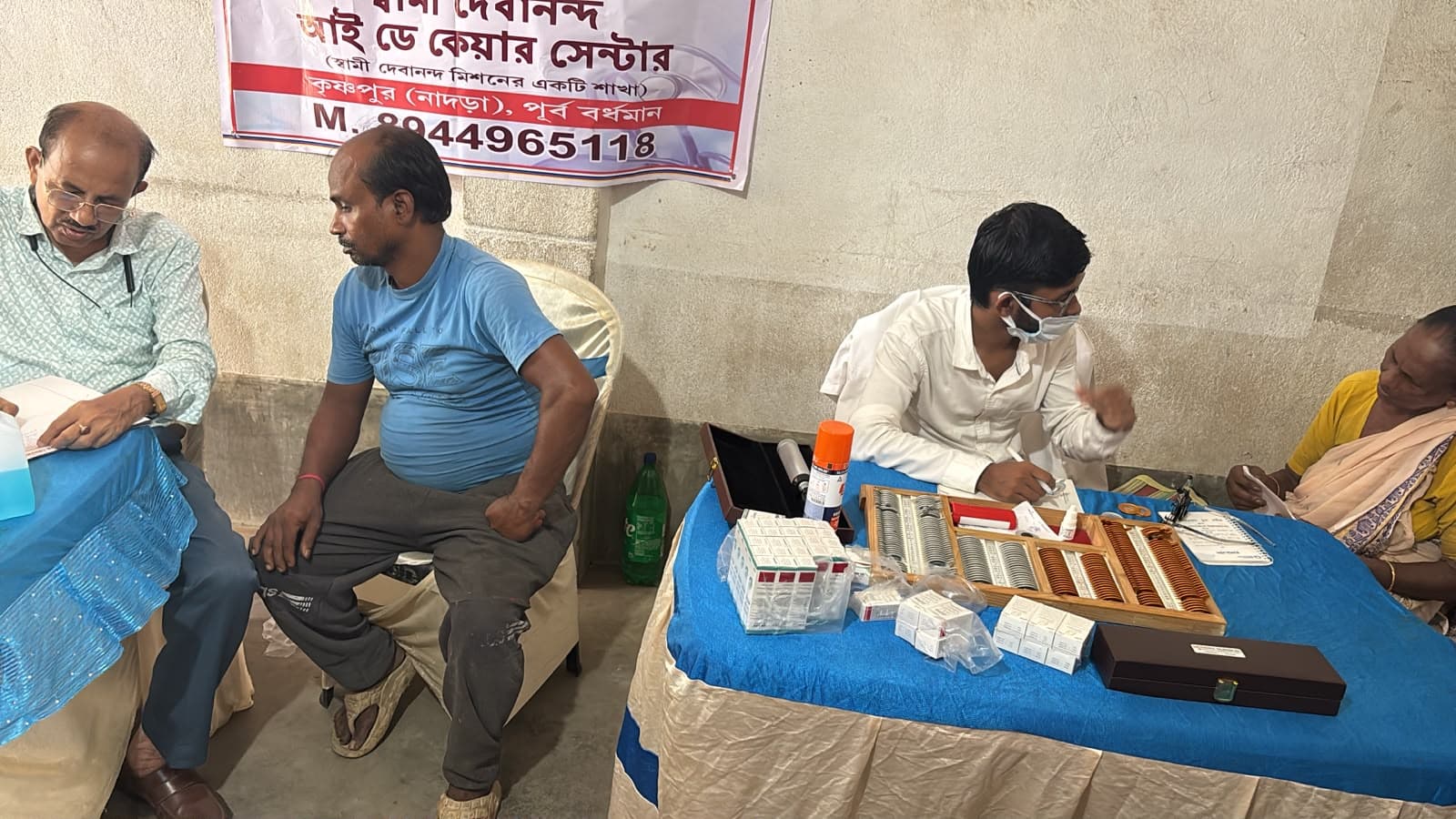An eye screening camp: a doctor seeing a patient at a table, with a banner lettered in Bengali behind