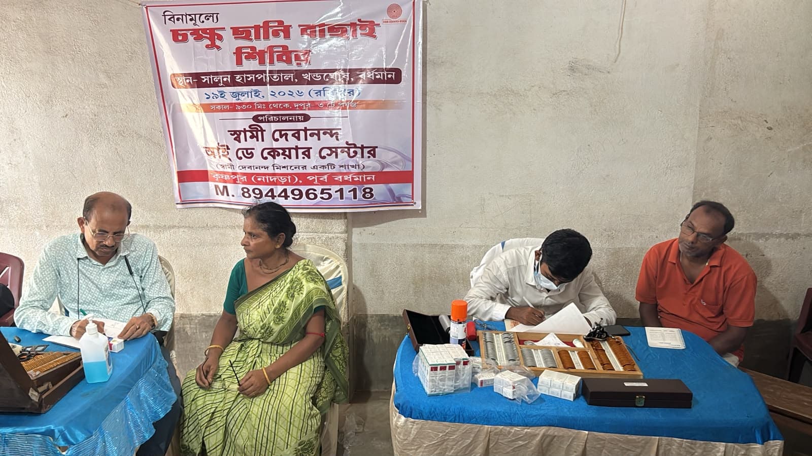 An eye screening camp: a doctor seeing a patient at a table, with a banner lettered in Bengali behind