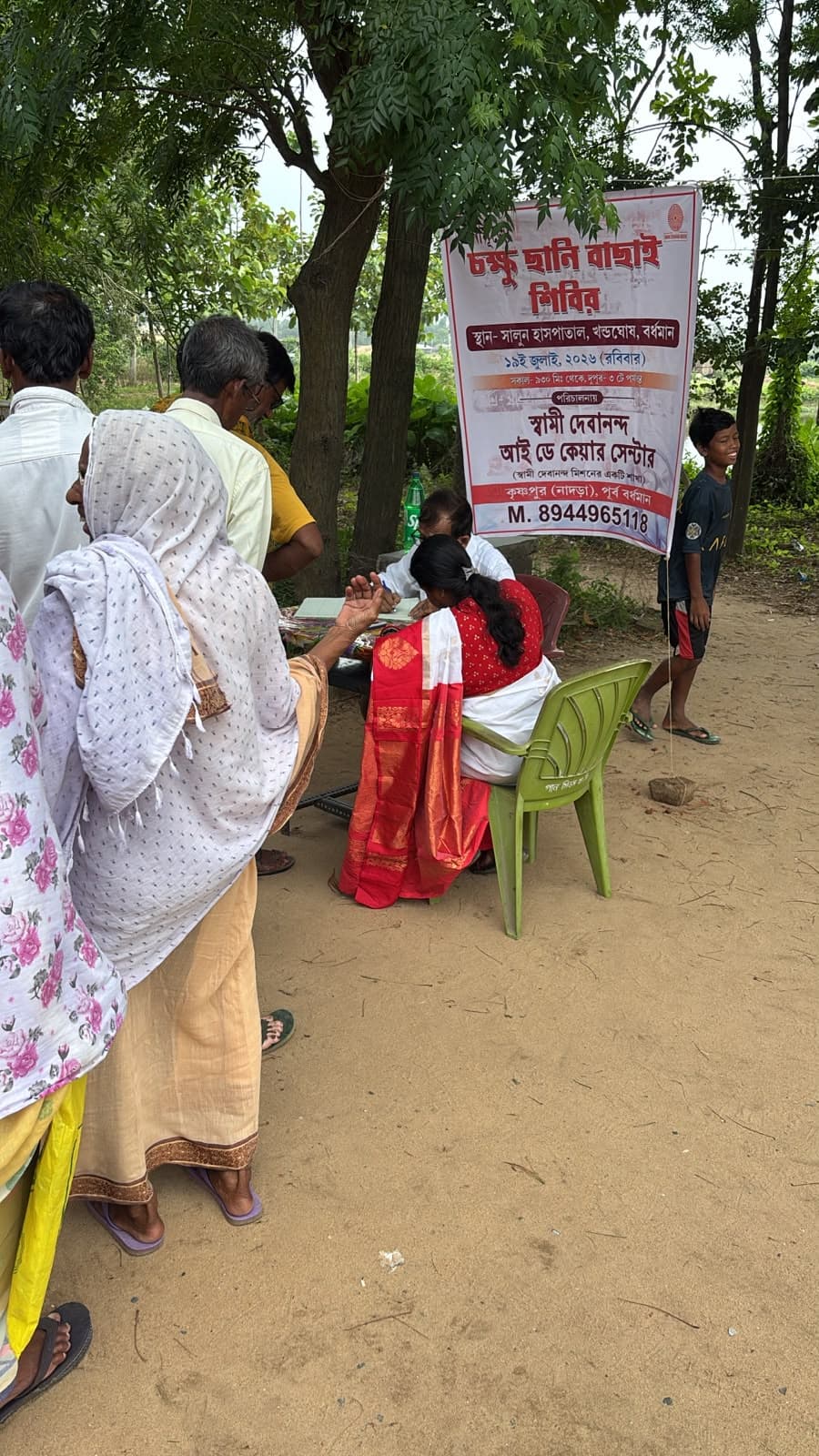 An eye screening camp: a doctor seeing a patient at a table, with a banner lettered in Bengali behind