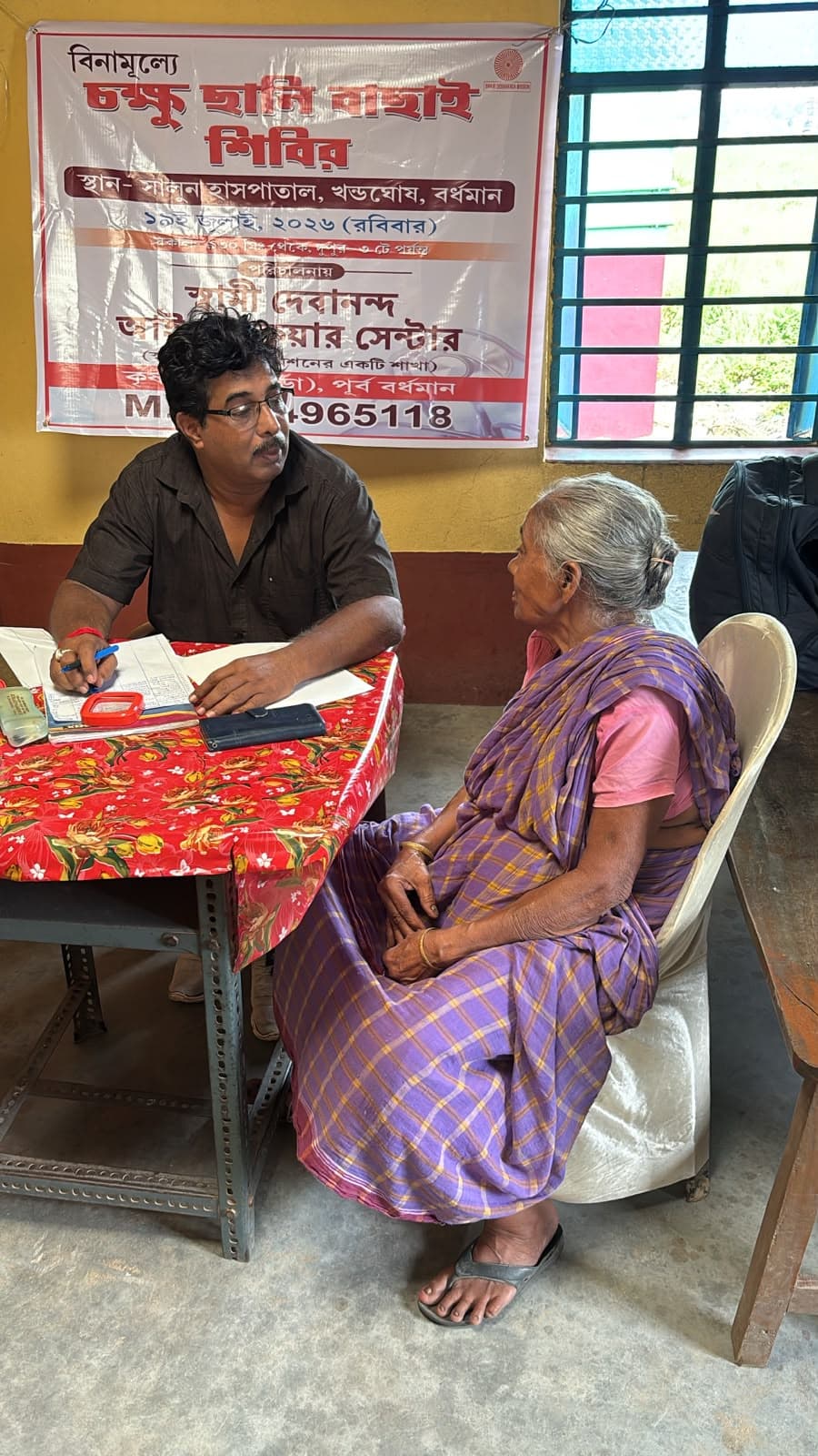 An eye screening camp: a doctor seeing a patient at a table, with a banner lettered in Bengali behind