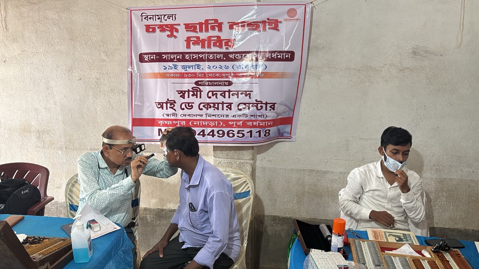 An eye screening camp: a doctor seeing a patient at a table, with a banner lettered in Bengali behind