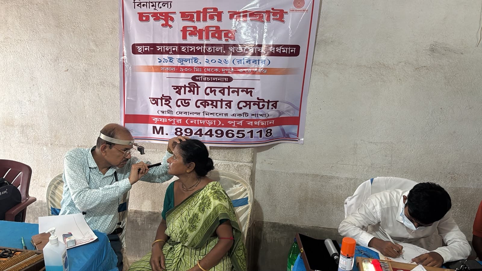 An eye screening camp: a doctor seeing a patient at a table, with a banner lettered in Bengali behind