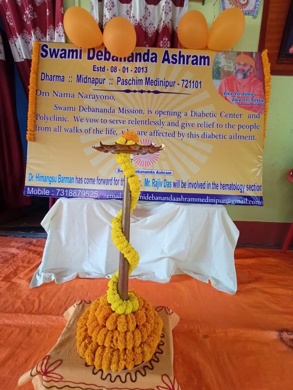 A brass lamp garlanded with marigolds in front of a banner of Swami Debananda Ashram, Dharma