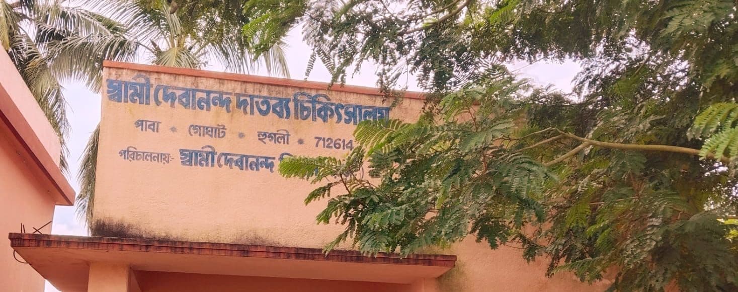 A single-storey dispensary building with its name painted in Bengali, beneath a spreading tree