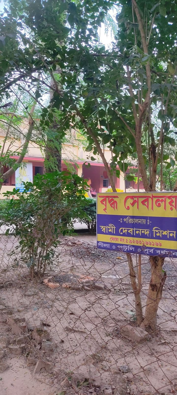 A signboard lettered in Bengali among young trees behind a wire fence