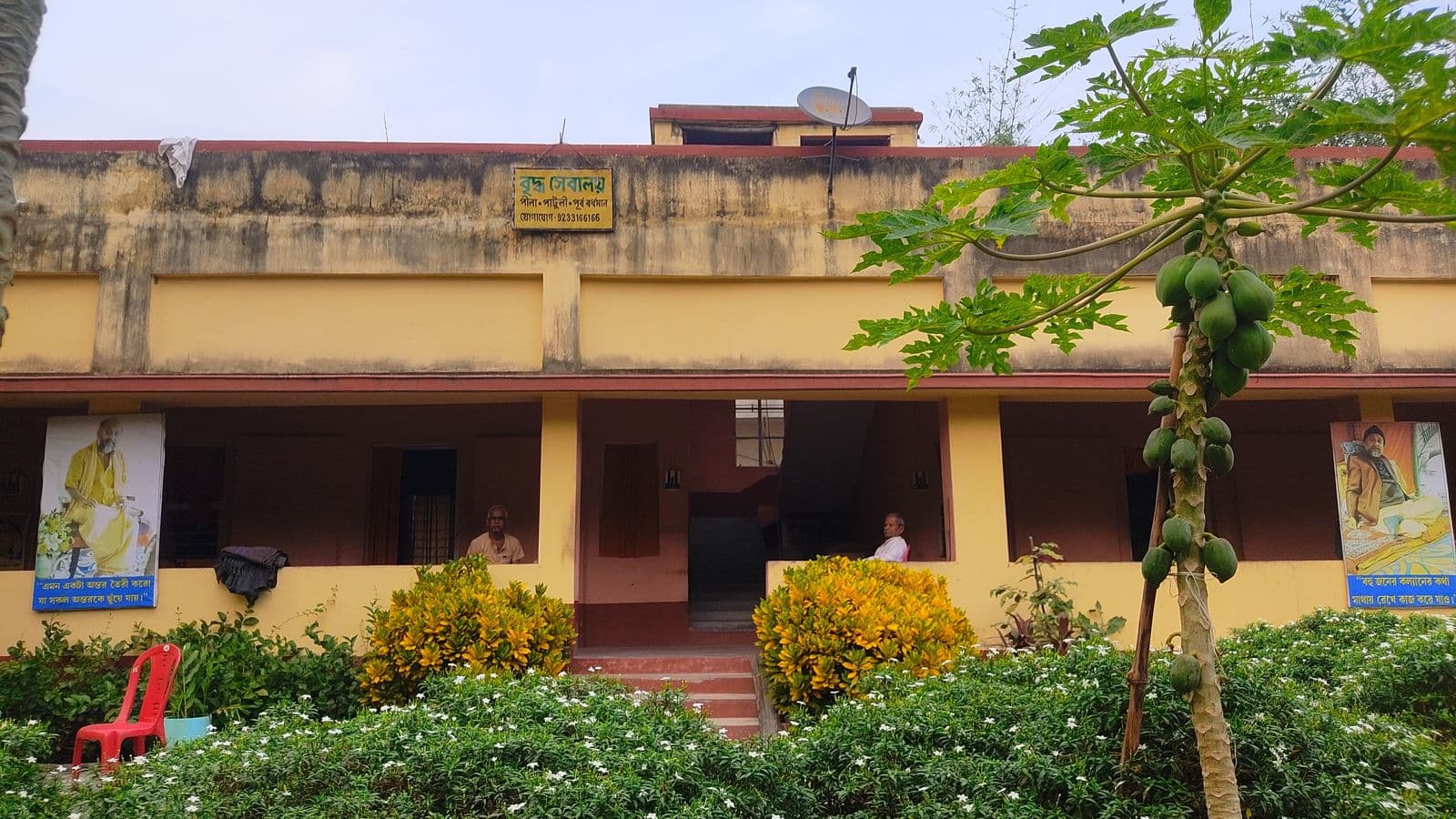 A two-storey building with a covered veranda behind flowering shrubs and a papaya tree