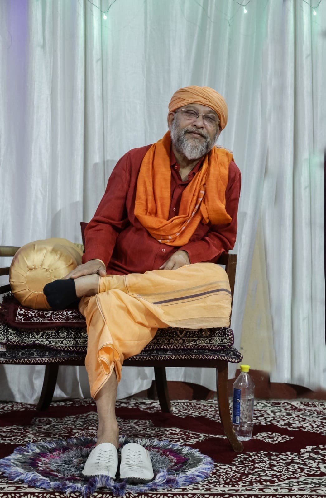 Swami Debananda Maharaj seated on a carved chair before white drapes, in a saffron turban and shawl