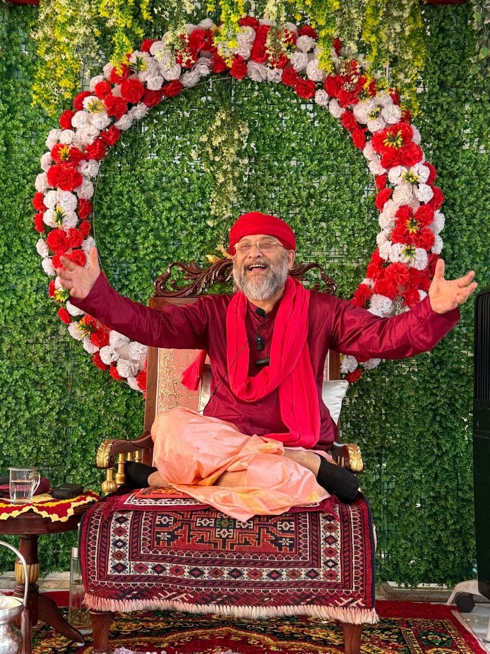 Swami Debananda Maharaj laughing with arms outstretched, seated before a ring of red and white flowers