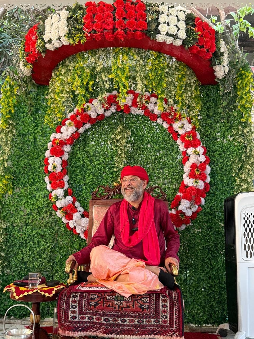Swami Debananda Maharaj seated on a carved chair before a wall of greenery and a ring of red and white flowers