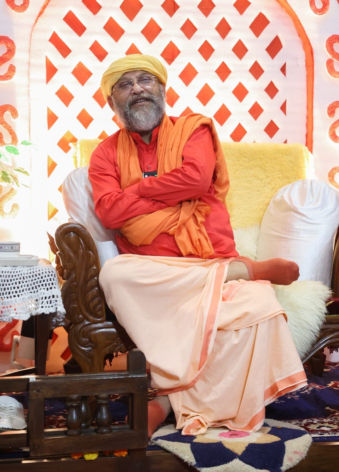 Swami Debananda Maharaj smiling, arms folded, seated before a lattice of orange diamonds
