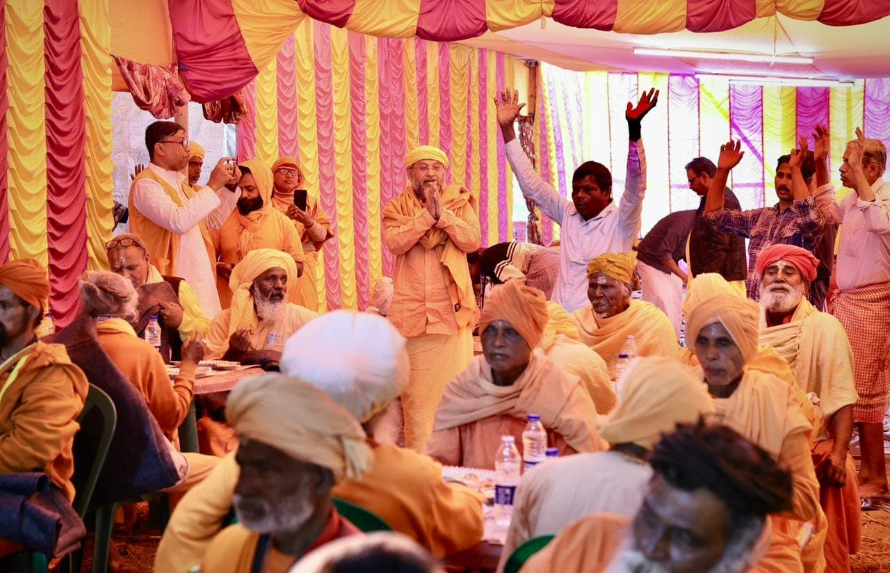 Swami Debananda Maharaj standing with palms joined among monks seated at tables under a striped canopy