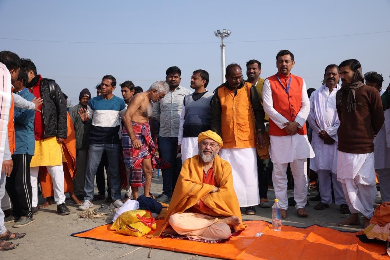 Swami Debananda Maharaj seated on a saffron cloth on open ground, surrounded by standing men