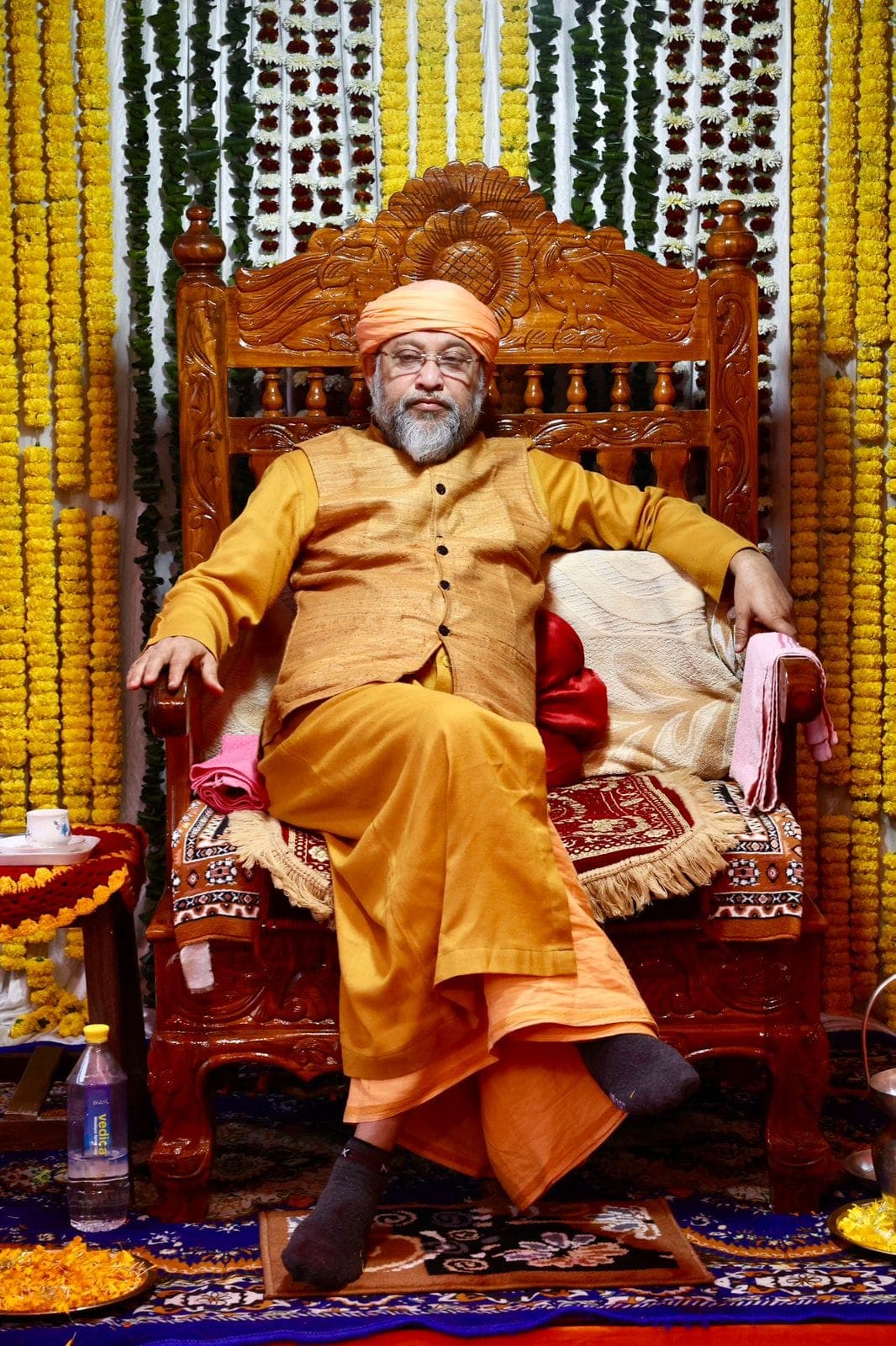 Swami Debananda Maharaj seated on a high carved chair framed by marigold garlands