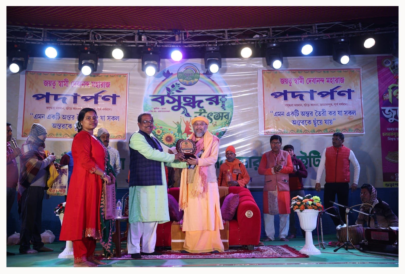 Swami Debananda Maharaj receiving a memento on a lit stage before banners in Bengali