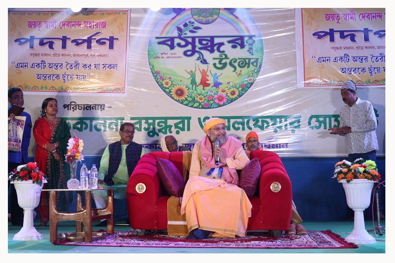 Swami Debananda Maharaj speaking into a microphone from a red sofa on a stage