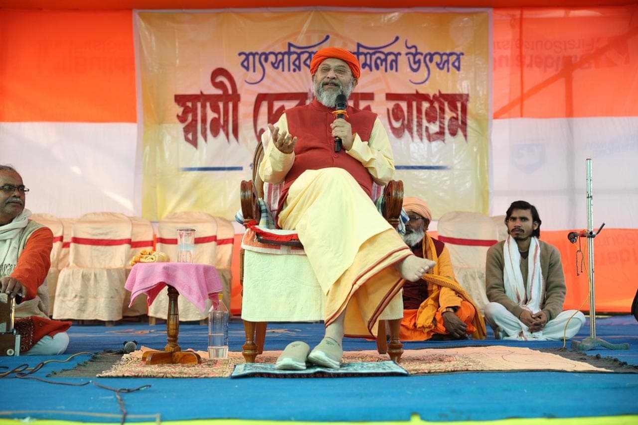 Swami Debananda Maharaj speaking into a microphone from a chair on a stage, with a banner in Bengali behind