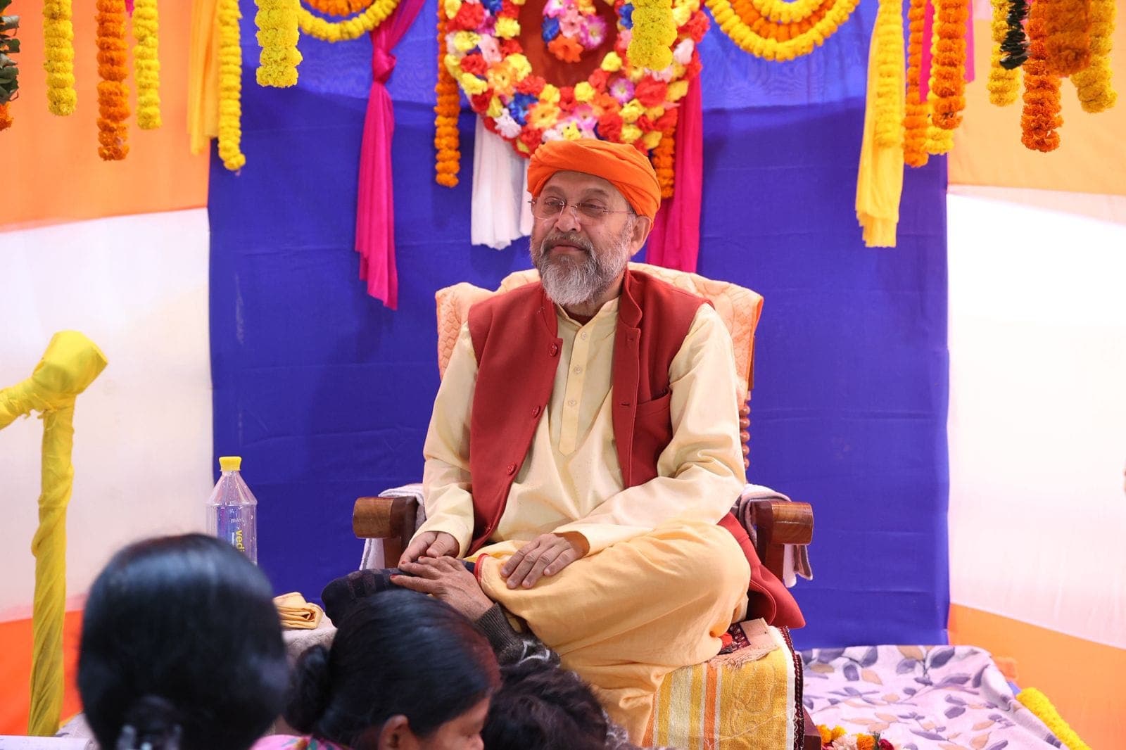 Swami Debananda Maharaj seated before a backdrop of marigold garlands, devotees seated in front