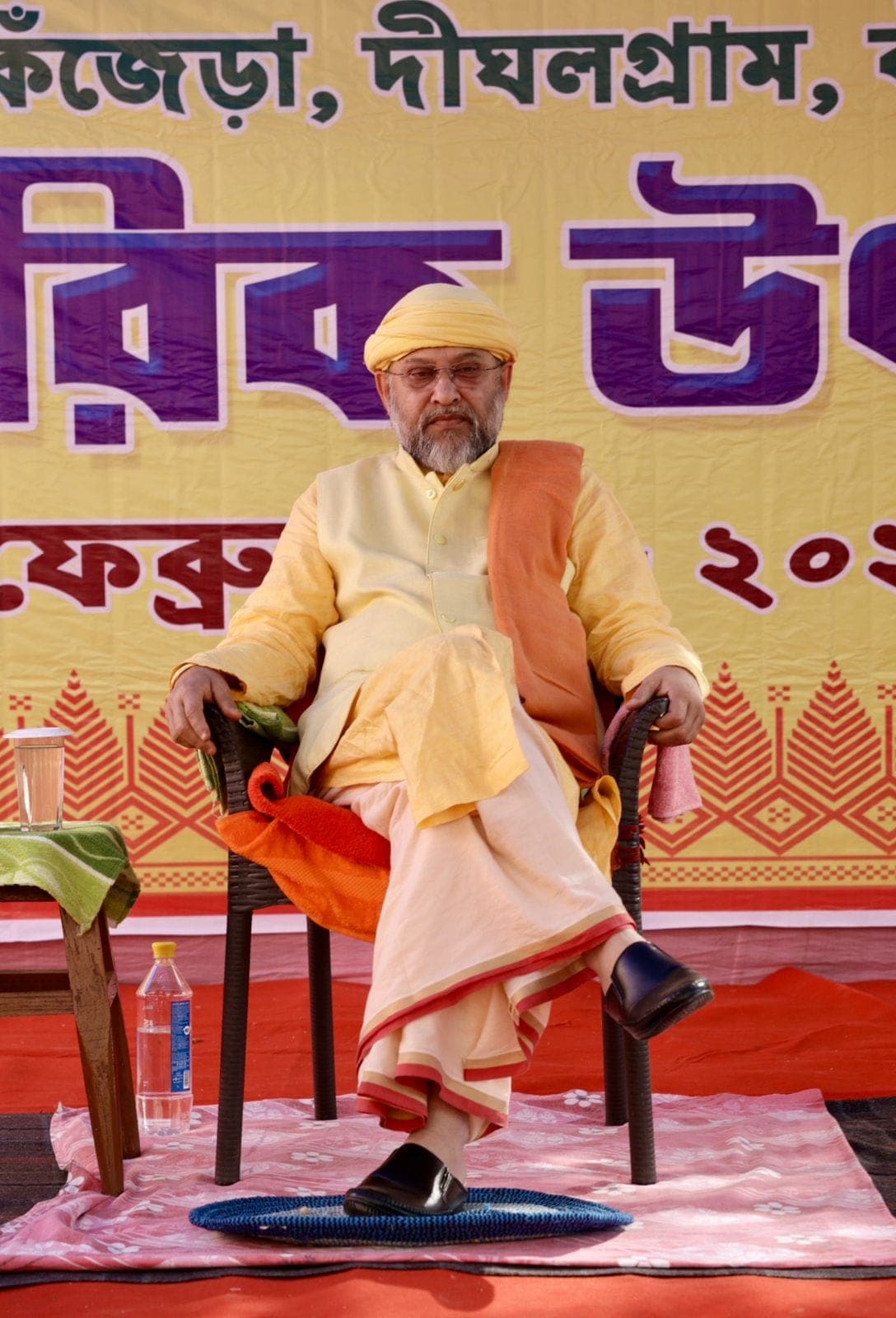 Swami Debananda Maharaj seated on a chair before a yellow banner lettered in Bengali
