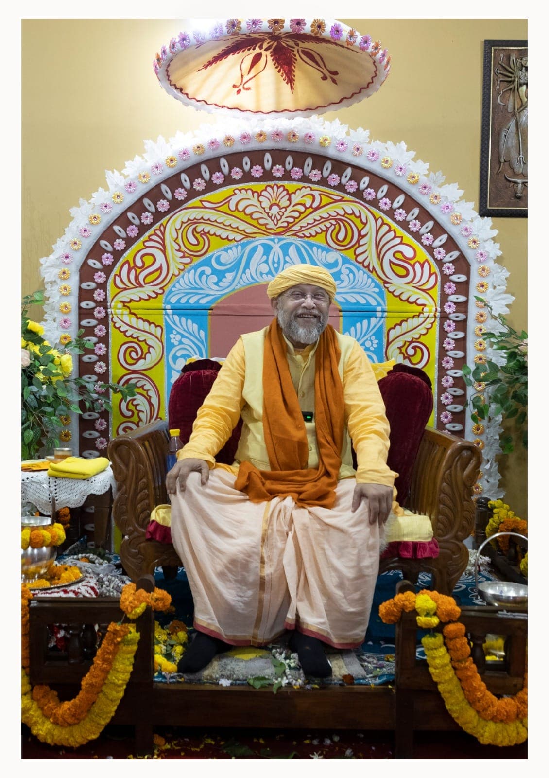 Swami Debananda Maharaj seated before a painted floral arch, with marigold garlands at his feet