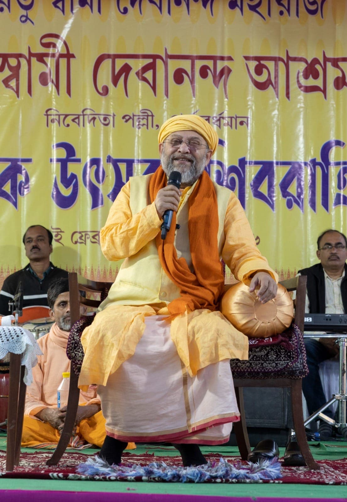 Swami Debananda Maharaj singing into a microphone, a drum in his other hand