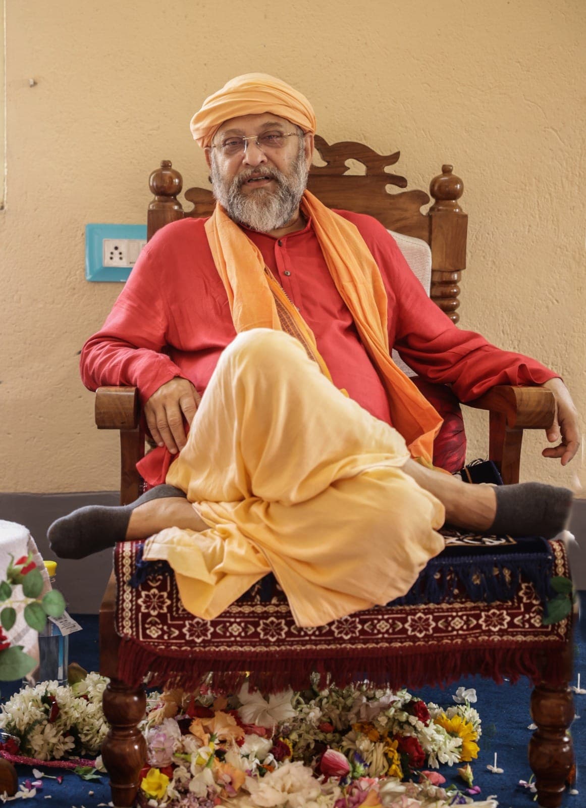 Swami Debananda Maharaj seated cross-legged on a carved chair, flowers strewn on the floor before him
