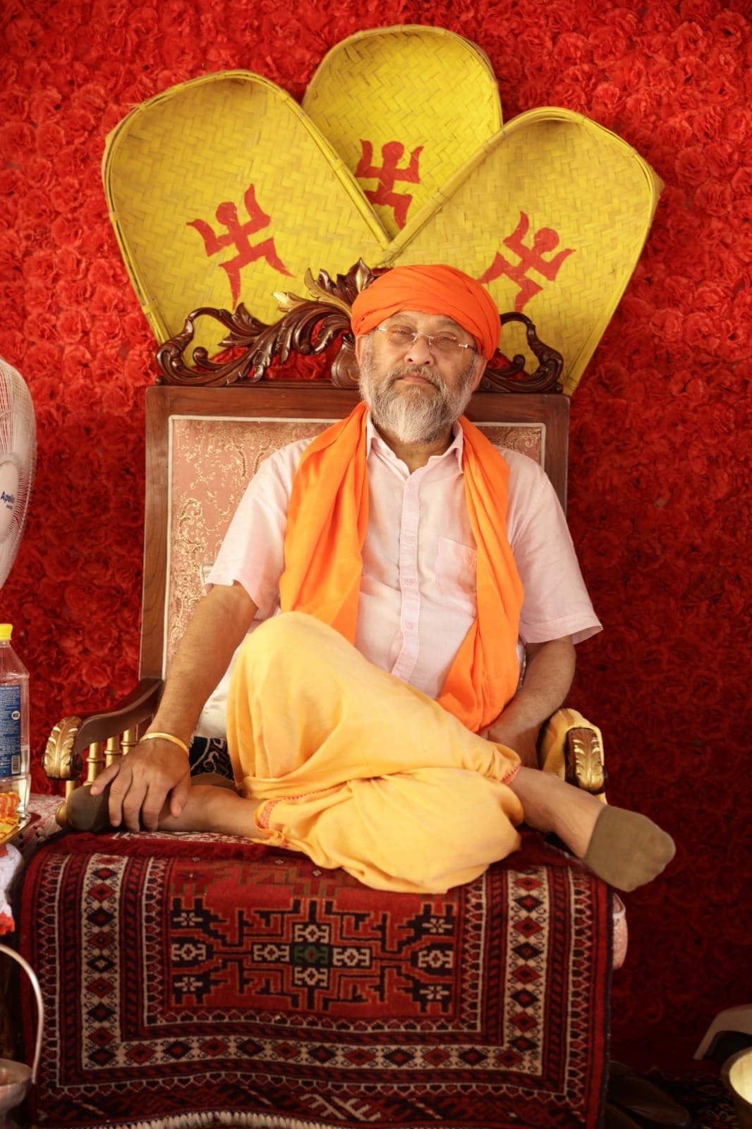 Swami Debananda Maharaj seated on a chair before a wall of red roses, in a saffron turban and shawl