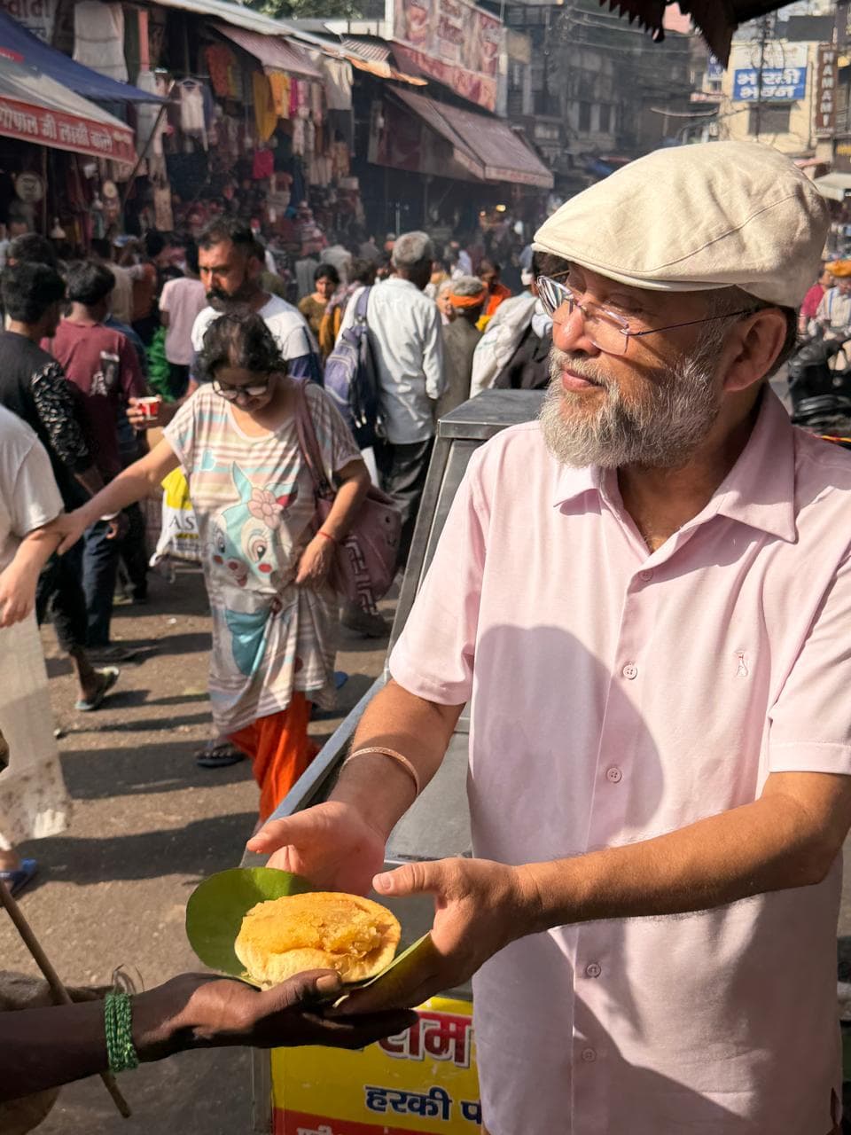 A man in a cap and pink shirt handing food on a leaf plate into an outstretched hand on a crowded street
