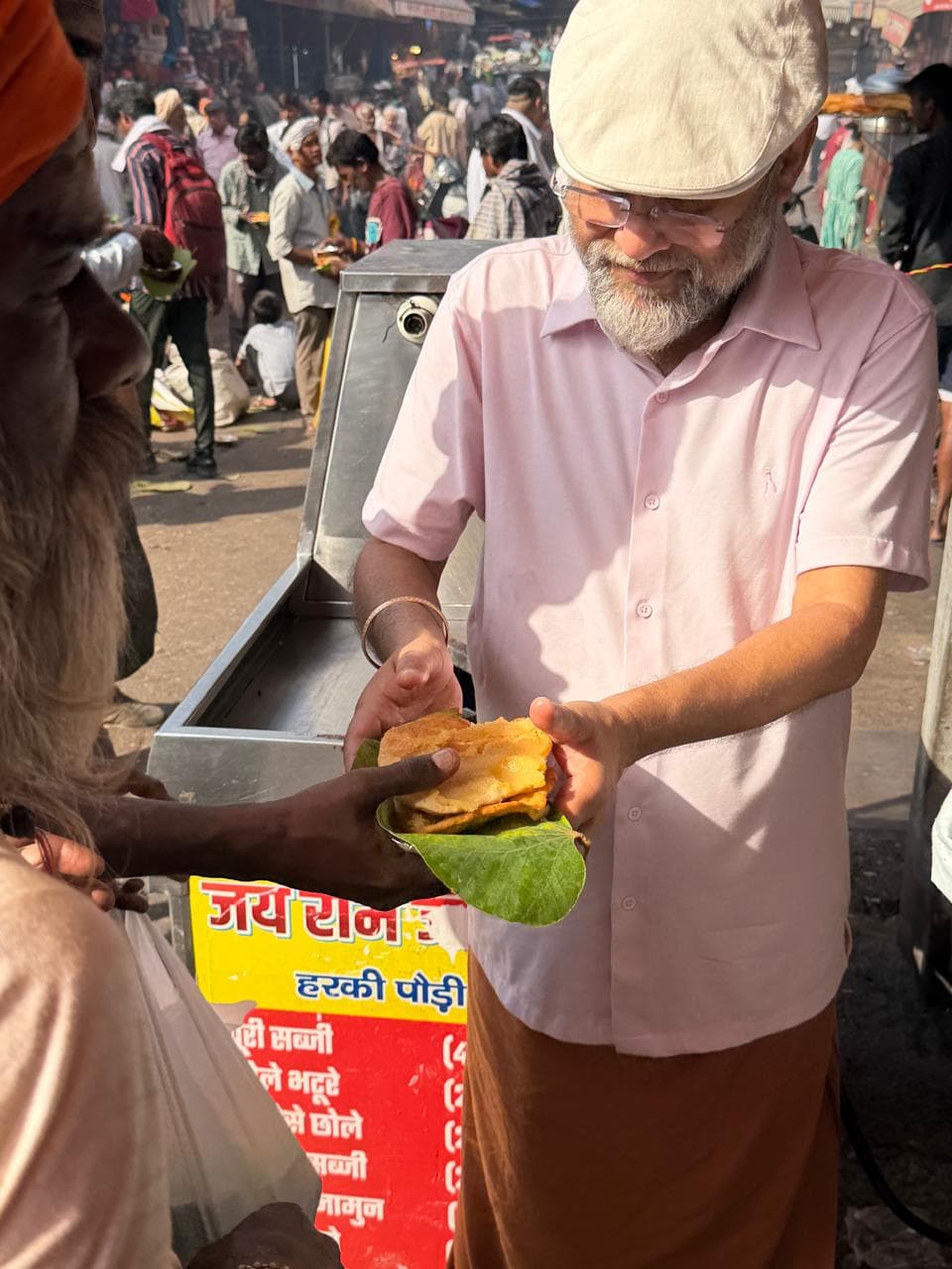 A man in a cap and pink shirt holding out food on a leaf plate, beside a sign lettered in Hindi