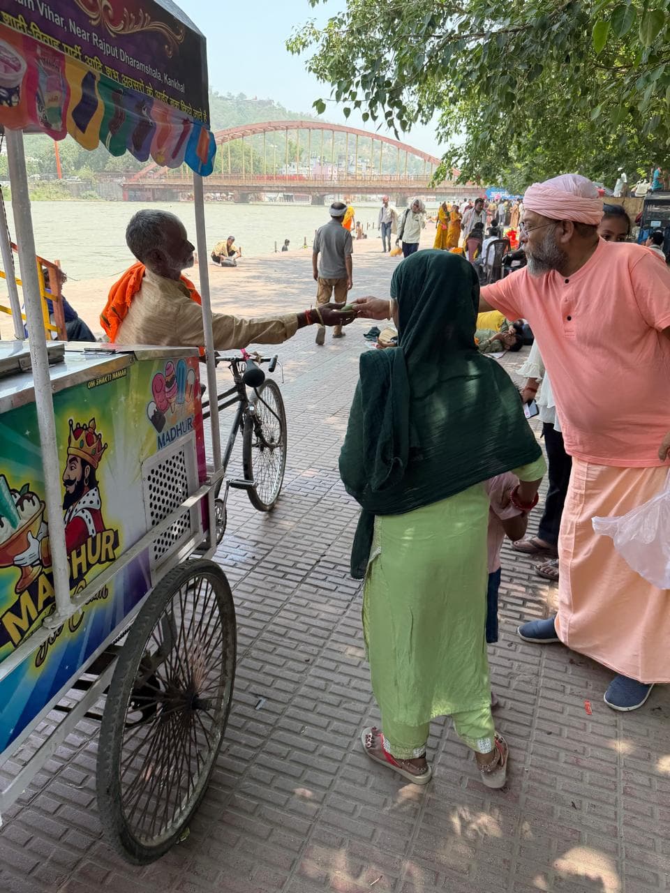 A man in saffron reaching out to a person beside a handcart on a riverside promenade, with a red bridge in the distance