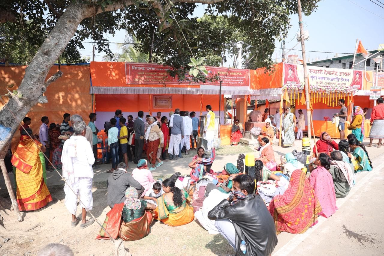 A crowd of people seated on the ground and standing in the shade of trees before an orange camp enclosure