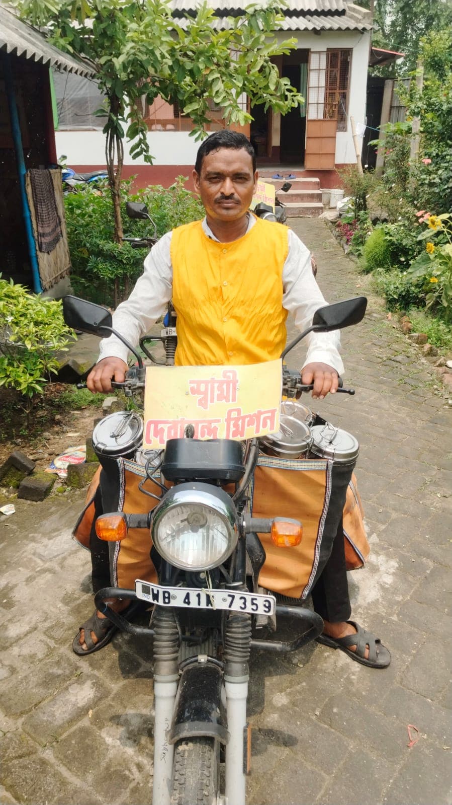 A man in a yellow vest seated on a motorcycle fitted with carrier bags, outside a house
