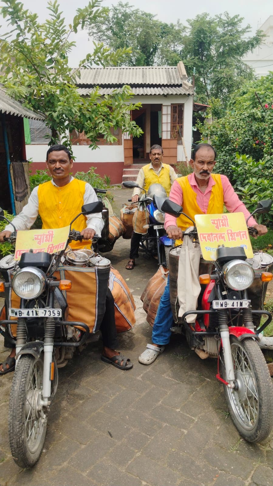 Two men in yellow vests on motorcycles fitted with carrier bags and Bengali signboards