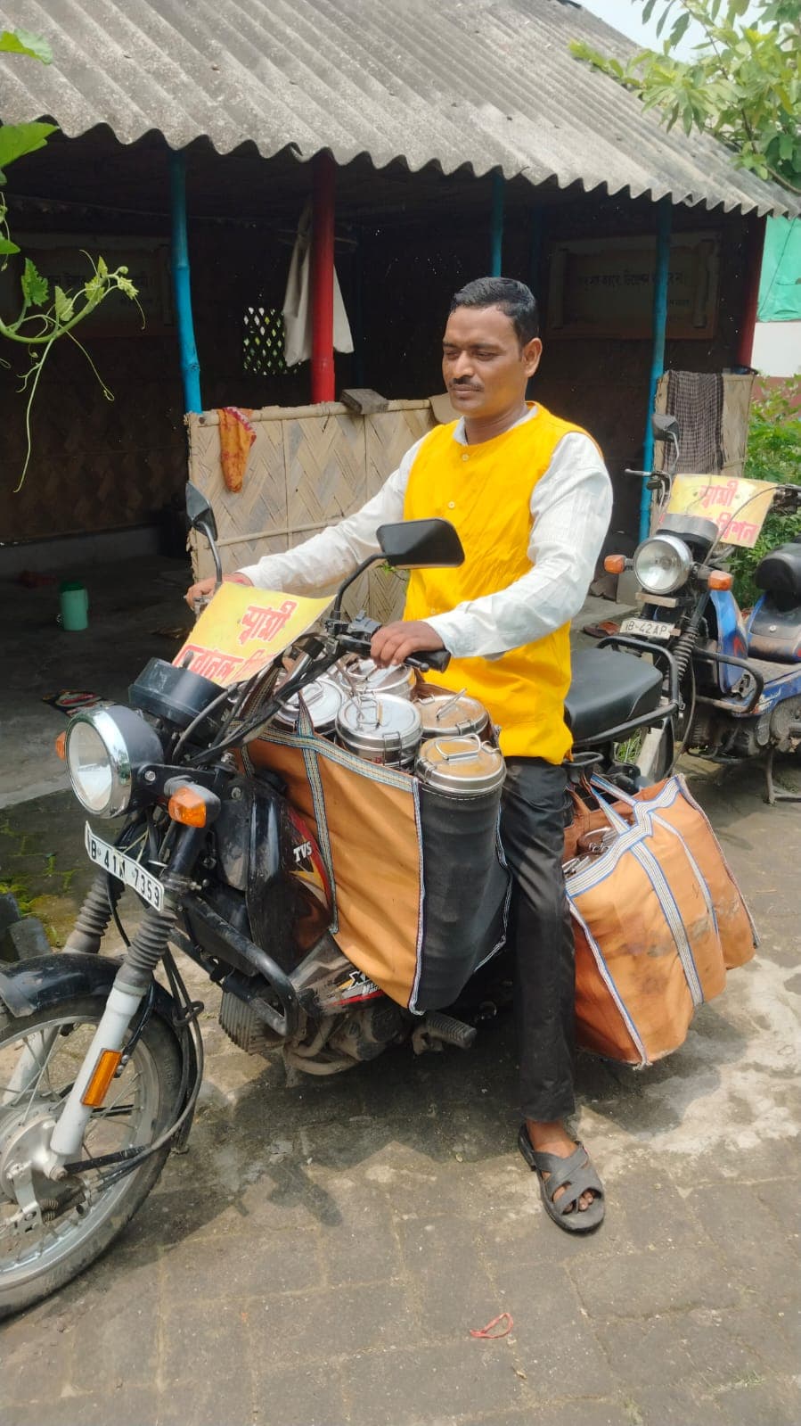 A man in a yellow vest on a motorcycle fitted with carrier bags, outside a house with a tin roof