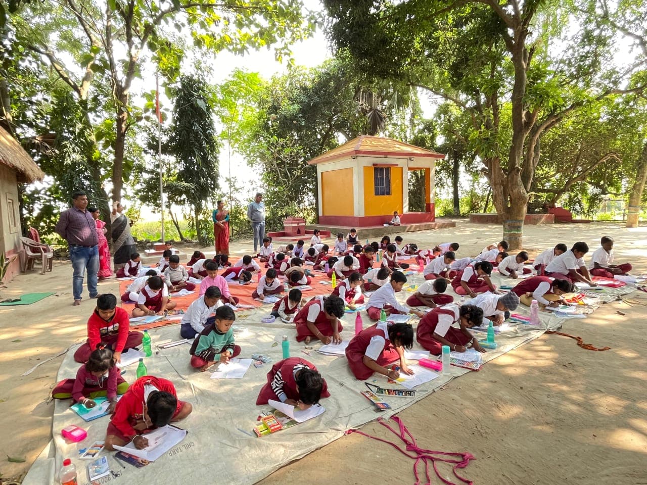 Students in uniform seated on mats under shade trees, bent over drawing paper, with a small yellow shrine behind them