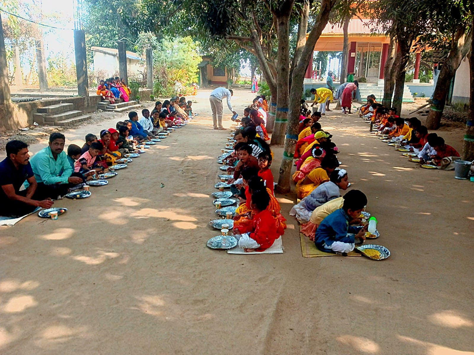Two long rows of children seated on mats along a sandy path under trees, with plates in front of them