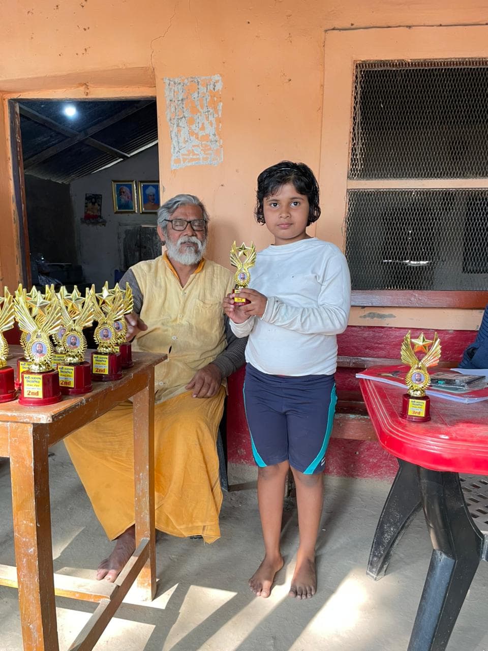 A young student holding a trophy beside a seated man in saffron, with more trophies on a table