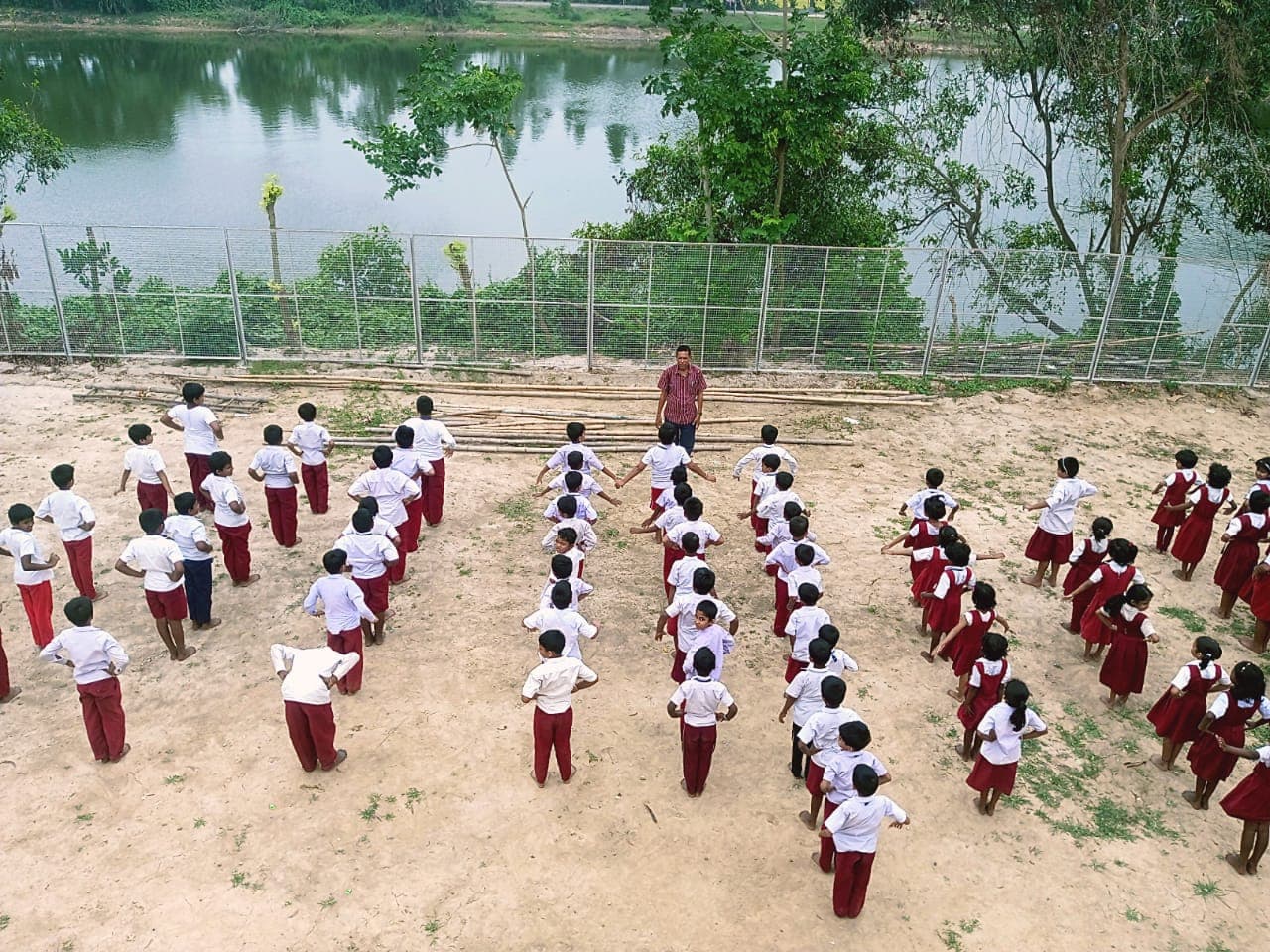 Rows of students in white and maroon uniforms doing exercises on a sandy ground beside a lake