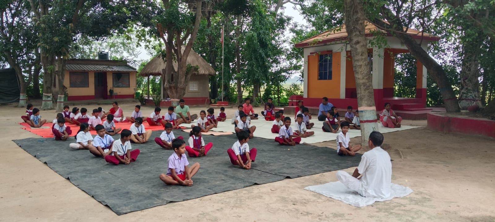 Students seated cross-legged on mats in a yard ringed by small buildings and trees, facing a teacher seated in white